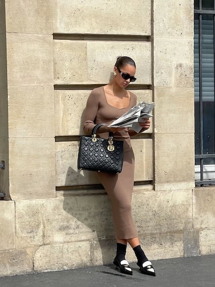 Woman in beige dress with Dior bag reading a newspaper by a stone wall.