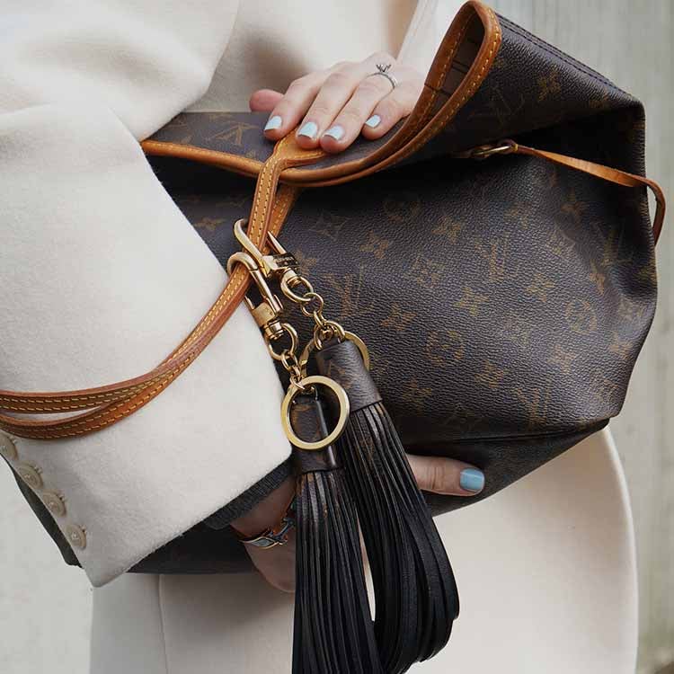 A Louis Vuitton Speedy 30 handbag in Damier Azur canvas with natural leather handles, adorned with a beige fur pom-pom and leather tassel charm, resting on a beige leather car seat.