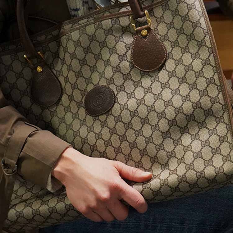 A close-up of a Gucci Horsebit 1955 shoulder bag in GG Supreme canvas with brown leather trim and gold horsebit hardware, resting on a person's lap dressed in white clothing with light pink manicured nails.