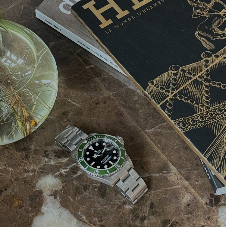 Rolex watch on marble table next to magazines and glass vase.