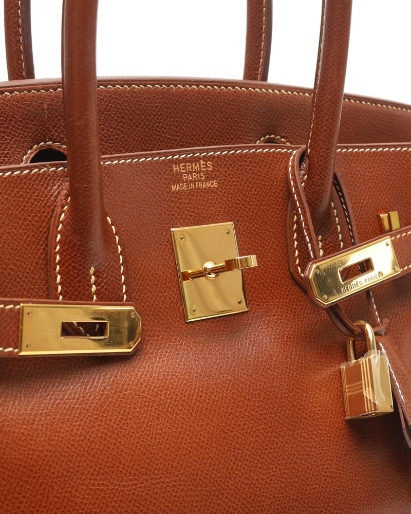 Schräge Nahaufnahme des goldfarbenen Verschlusses, Schlosses und Markenstempels der Hermès Birkin 30 auf braunem Epsom-Leder.
