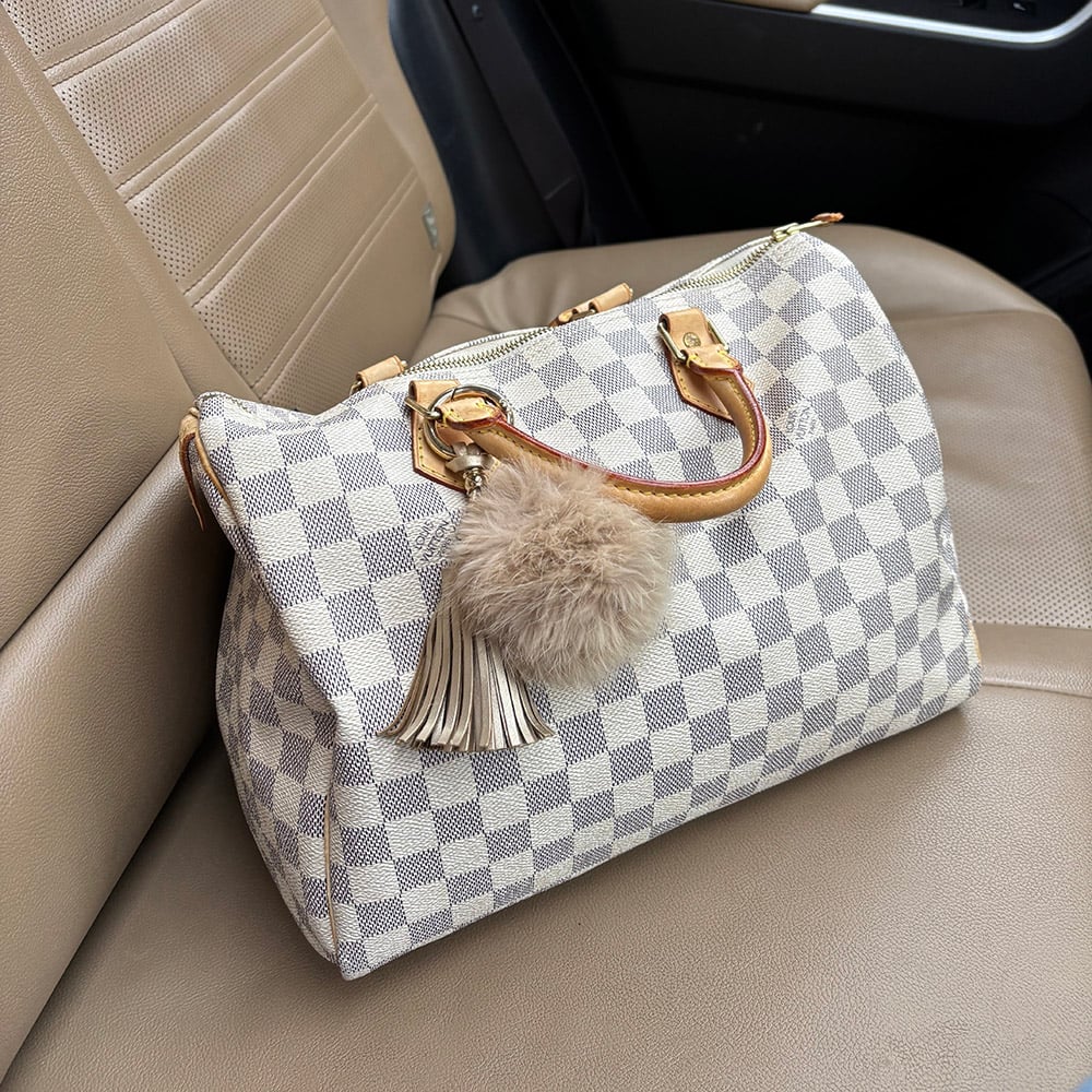 A Louis Vuitton Speedy 30 handbag in Damier Azur canvas with natural leather handles, adorned with a beige fur pom-pom and leather tassel charm, resting on a beige leather car seat.