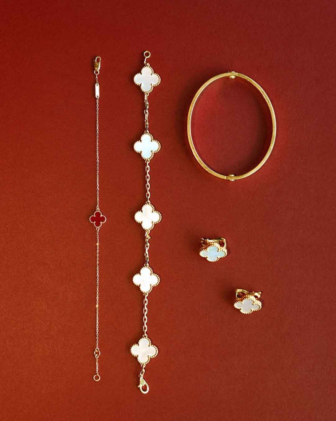 A set of elegant gold and mother-of-pearl jewelry displayed on a deep red background, including bracelets, earrings, and a bangle in a clover-shaped design.