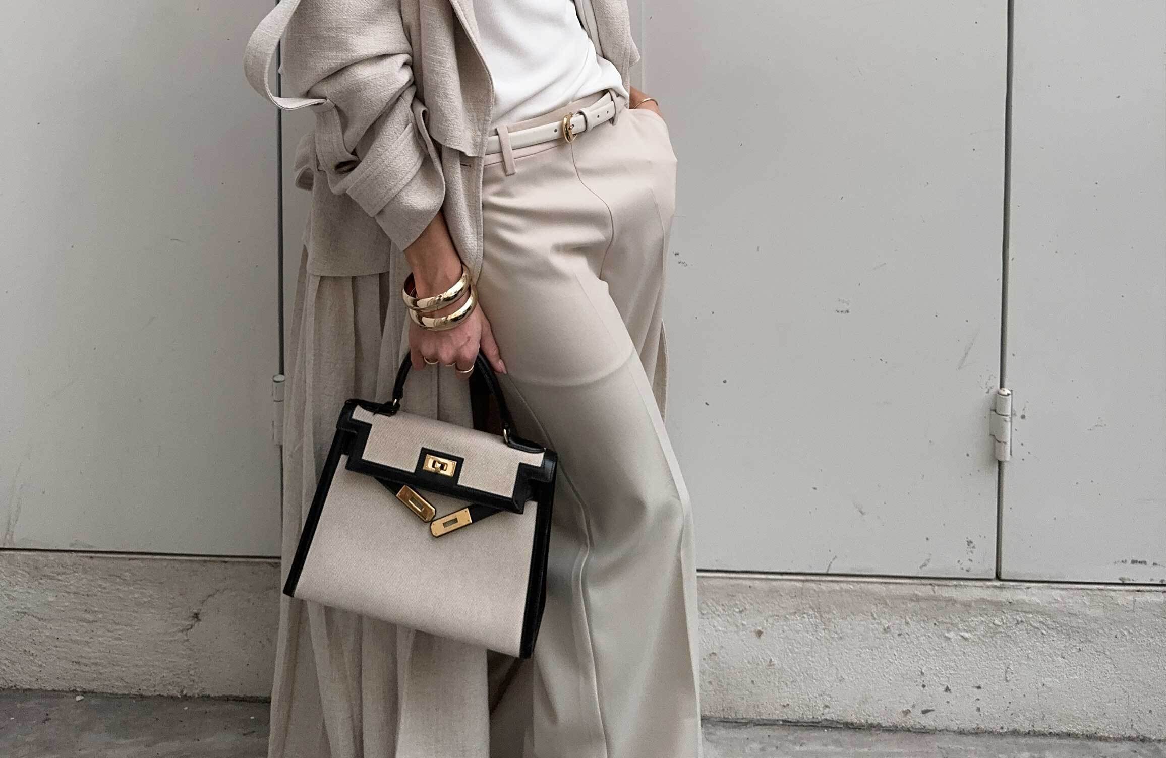 Person in beige outfit holding a beige Hermès Kelly bag with black details.