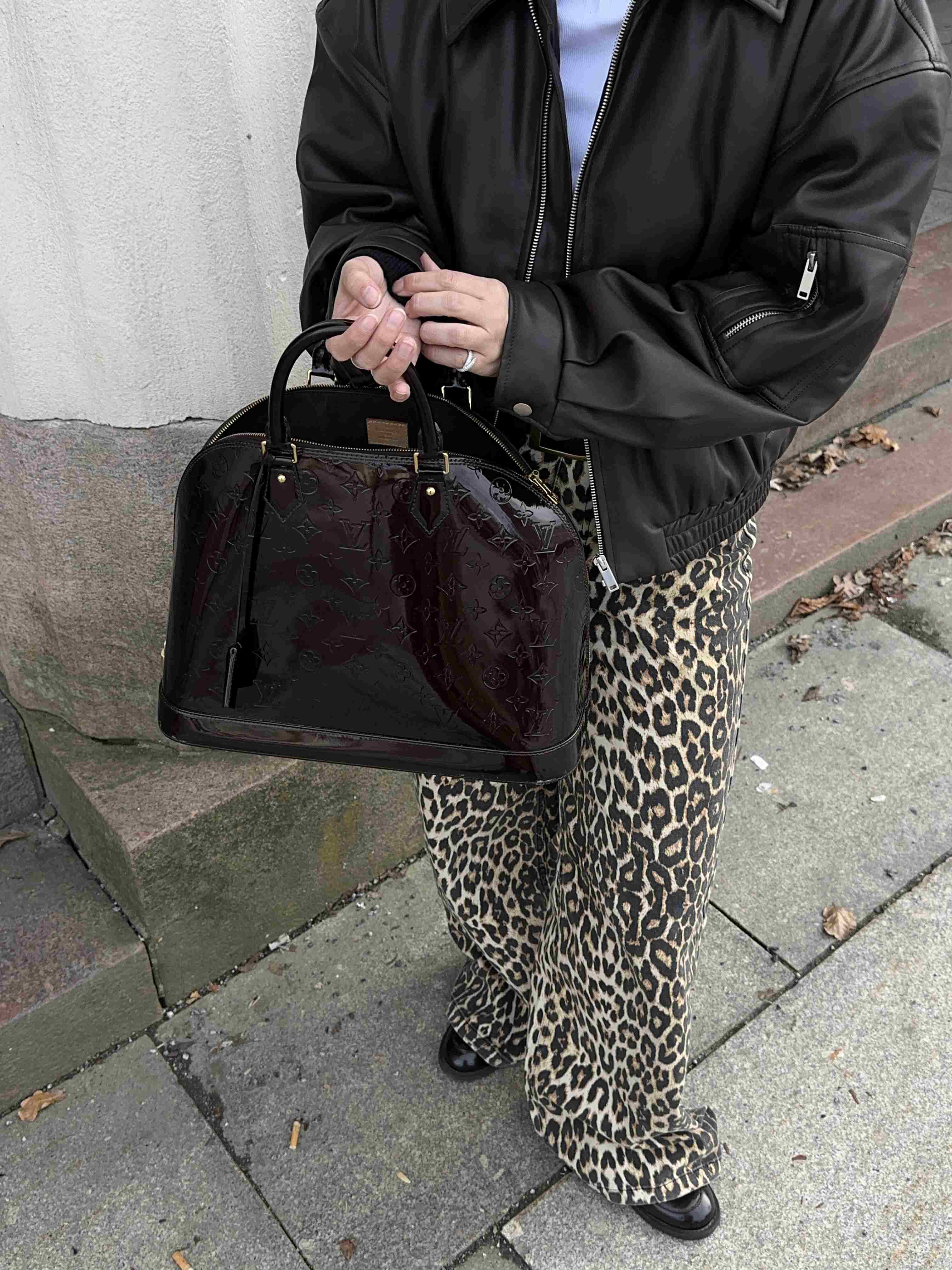 Shiny dark Louis Vuitton bag held by a person in leather jacket and leopard pants.