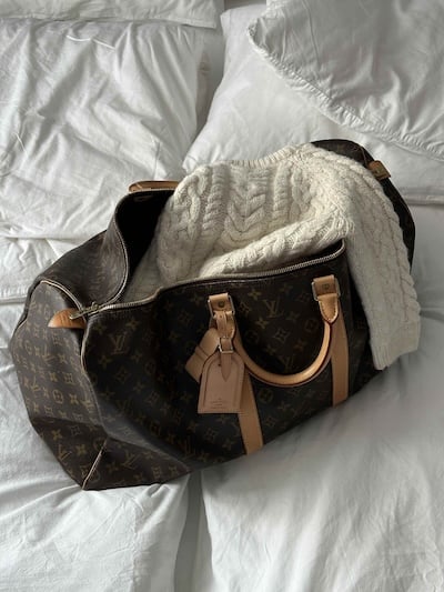 Open Louis Vuitton travel bag with white knitted sweater on a bed.