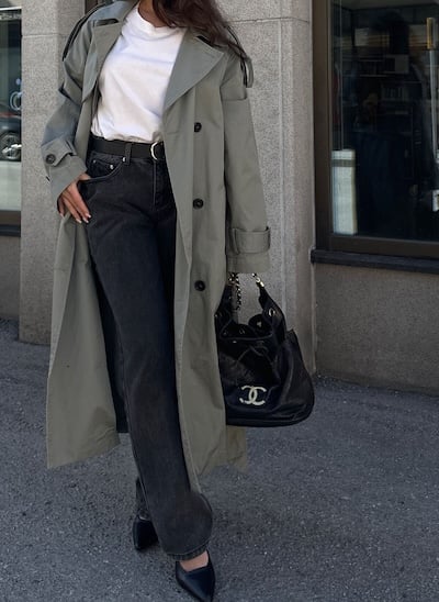 Woman in trench coat with black Chanel bag and black jeans on sidewalk.