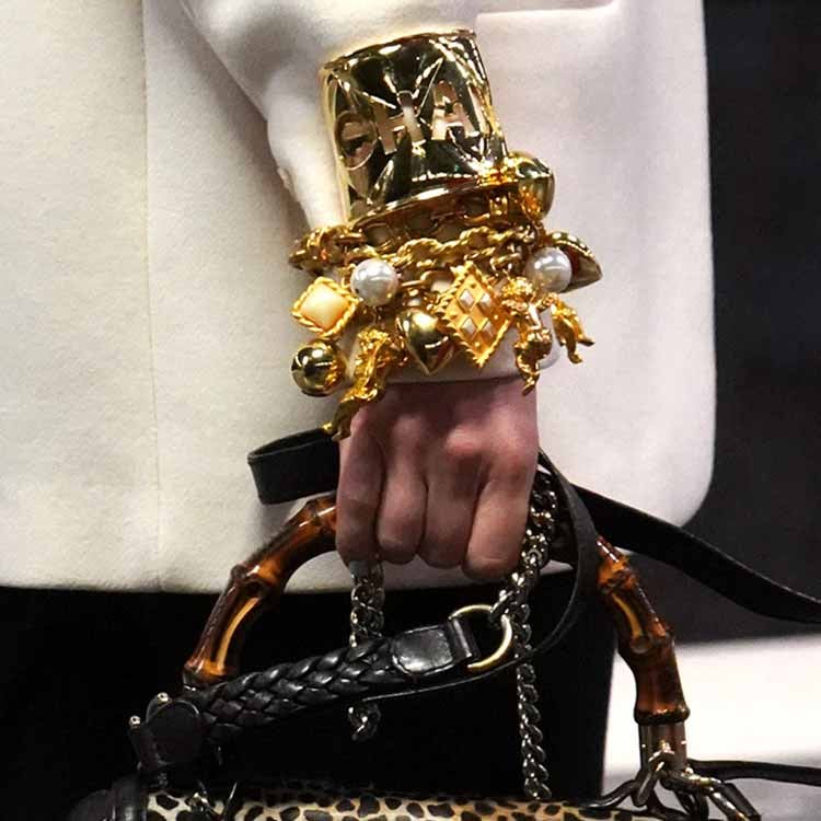 A stylish woman with long blonde hair, wearing sunglasses and a brown outfit, stands at a Parisian-style newsstand reading a magazine. She carries a textured brown shoulder bag, while the stand around her is filled with colorful newspapers and magazines.